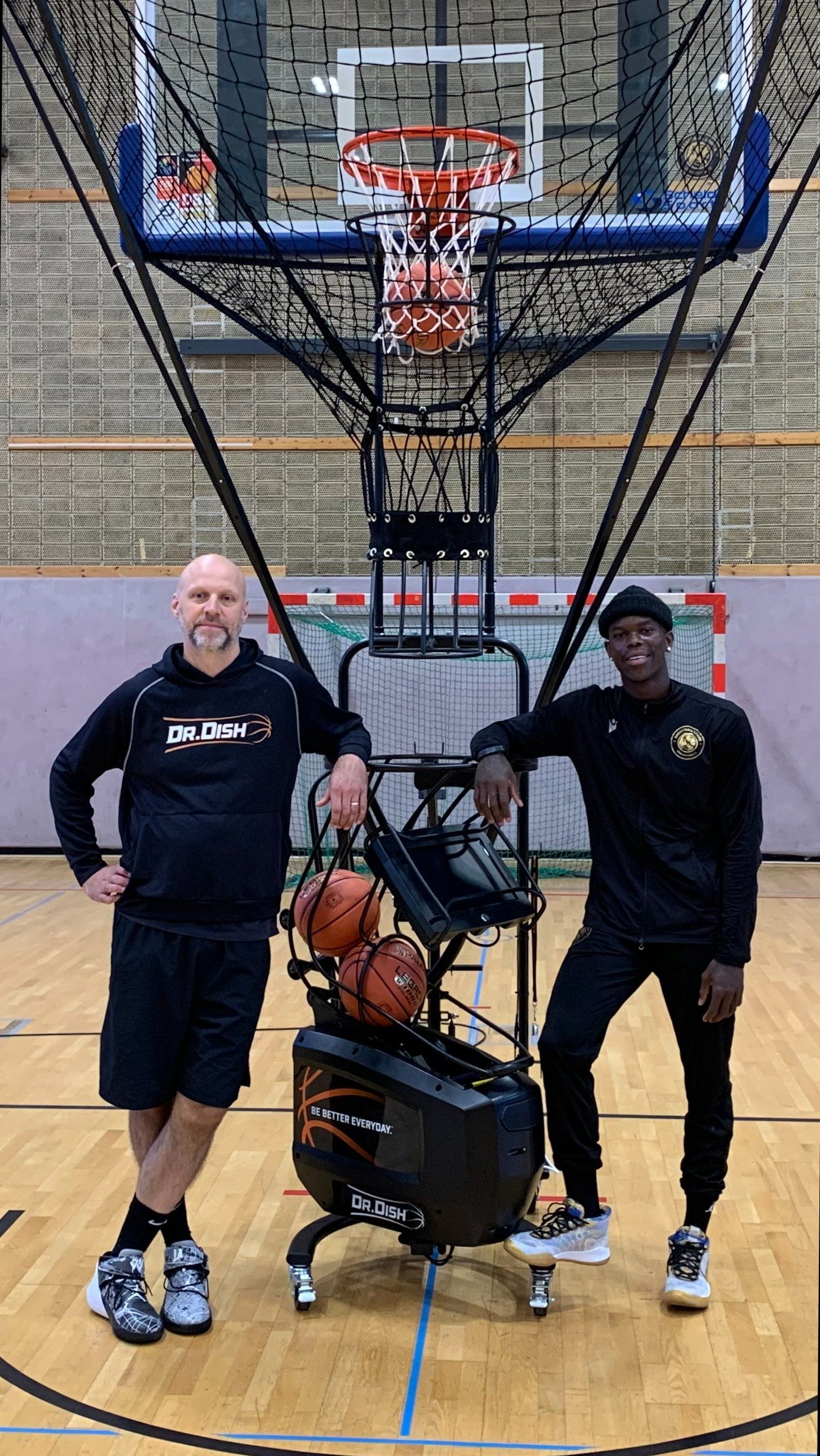 Dennis Schröder standing with the Dr. Dish CT+ basketball shooting machine at Basketball Löwen Braunschweig's training facility in Germany.